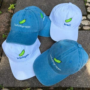 Könnte beinhalten: Vier Baseballkappen in Blau und Weiß. Zwei blaue Kappen zeigen gestickte grüne Gurken und den Text 'tzatzikigruppe'. Eine weiße Kappe zeigt eine grüne Gurke und 'braut', eine weitere weiße Kappe zeigt eine grüne Gurke und 'bräutigam'.