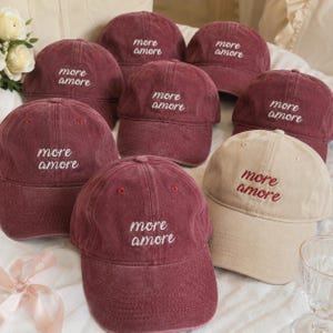 Könnte beinhalten: Eine Auswahl an Baseballkappen in verschiedenen Burgunderfarben und eine beige. Jede Kappe ist mit dem Schriftzug "more amore" in weißer oder roter Farbe bestickt. Die Kappen liegen auf einer weißen Oberfläche.