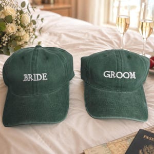 May include: Two dark green baseball caps with structured crowns and curved brims. The left cap features the word BRIDE embroidered in white thread, while the right cap features the word GROOM embroidered in white thread on the front panels.