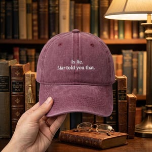 Può includere: Cappellino da baseball bordeaux con la scritta bianca "Is lie. Liar told you that.". Il cappellino è tenuto davanti a una libreria piena di libri.
