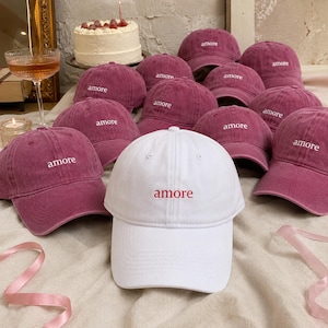 Puede incluir: Una colección de gorras de béisbol, principalmente en un tono rojo oscuro, con la palabra "amore" bordada en blanco o rojo. Destaca una gorra blanca con bordado rojo. Las gorras están dispuestas sobre una superficie clara.