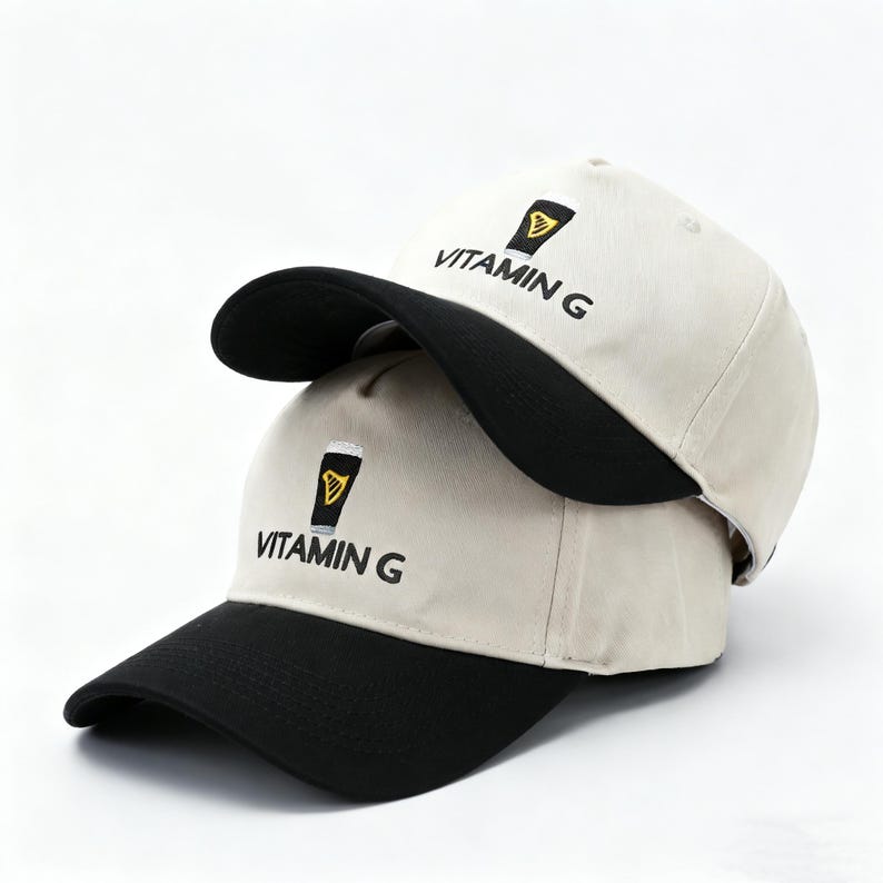 May include: Two beige baseball caps with black brims are stacked. Each cap features the embroidered text "VITAMIN G" below a small, stylized graphic of a pint of beer. The caps are made of a soft fabric.