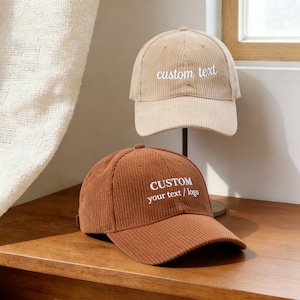 Könnte beinhalten: Zwei Cord-Baseballkappen, eine beige und eine braune, mit gebogenen Schirmen. Auf der beigefarbenen Kappe ist "custom text" auf der Vorderseite gestickt. Auf der braunen Kappe steht "CUSTOM your text / logo". Beide Kappen liegen auf einer Holzoberfläche.
