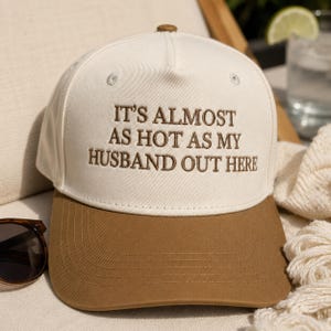 Könnte beinhalten: Eine cremefarbene und braune Baseballkappe mit dem gestickten Text "IT'S ALMOST AS HOT AS MY HUSBAND OUT HERE". Die Kappe hat einen gebogenen Schirm und einen Knopf oben. Sonnenbrillen sind im Hintergrund zu sehen.