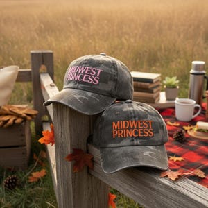 Könnte beinhalten: Zwei graue Camouflage-Baseballkappen mit dem Text "MIDWEST PRINCESS" in Pink und Orange. Die Kappen liegen auf einem Holzzaun, mit einer Picknick-Szene im Hintergrund.