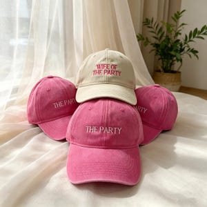 Può includere: Un gruppo di cappellini da baseball rosa e beige. I cappellini rosa presentano la scritta bianca "THE PARTY", mentre quello beige ha la scritta rosa "WIFE OF THE PARTY". I cappellini sono in tessuto morbido e strutturato.