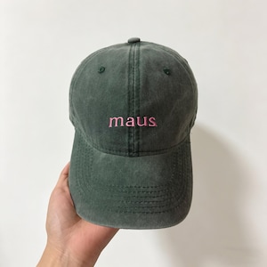 Könnte beinhalten: Dunkelgrüne Baseballkappe mit dem Wort "maus" in Rosa gestickt. Die Kappe hat einen gebogenen Schirm und einen Knopf oben. Der Hut ist aus einem weichen, abgenutzten Stoff gefertigt.