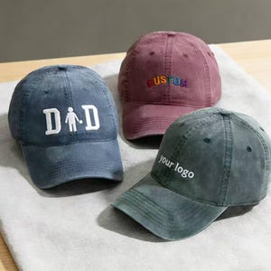 Könnte beinhalten: Drei Baseballkappen in Blau, Burgunderrot und Grün. Die blaue Kappe hat das Wort "DAD" mit einer Strichmännchen-Figur. Die bordeauxrote Kappe trägt das Wort "CUSTOM" in Regenbogenfarben. Die grüne Kappe hat den Text "your logo" in Weiß.