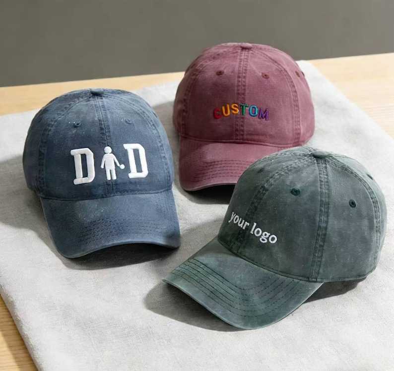 May include: Three baseball caps in blue, burgundy, and green. The blue cap features the word "DAD" with a stick figure. The burgundy cap has the word "CUSTOM" in rainbow colours. The green cap has the text "your logo" in white.