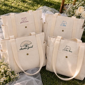 May include: Four off-white canvas tote bags with dual handles, side pockets, and a front pocket secured by a brass snap. Each bag features unique embroidery: an envelope with 'girl mom', a blue dolphin with 'Lea', a fan with 'chaotic good', and a dolphin with 'Ann-Marie'.