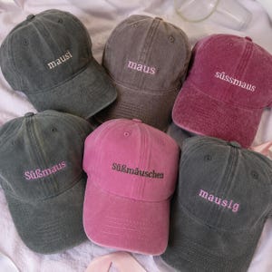 Könnte beinhalten: Sechs Baseballkappen in Anthrazit, Braun und Magenta, jede mit gesticktem Text auf der Vorderseite. Die Kappen zeigen die Wörter mausi, maus, süssmaus, Süßmaus, Süßmäuschen und mausig in verschiedenen Garnfarben, darunter Weiß, Rosa und Schwarz.