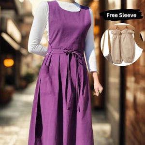 以下が含まれることがあります： ノースリーブの紫色のエプロンドレス。ウエストにリボン、ポケット付き。白い長袖シャツの上に重ね着されています。画像には、ベージュの袖カバーが2つと「Free Sleeve」というテキストも含まれています。