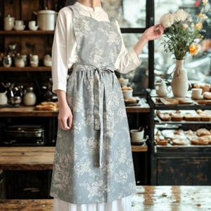 May include: A gray apron with a white floral pattern. The apron has a bib, waist tie, and a full skirt. The apron is worn over a white blouse and skirt. The background shows a kitchen setting with baked goods and flowers.