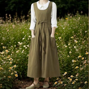 Könnte beinhalten: Olivgrünes Latzkleid mit Bindegürtel und großen Taschen. Das Kleid wird über einer weißen Bluse mit rundem Kragen getragen. Das Model trägt passende grüne Schuhe. Der Hintergrund ist ein Blumenfeld.