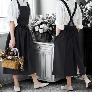Puede incluir: Delantal negro con cuello cuadrado y falda larga. El delantal tiene una atadura en la cintura y bolsillos. El modelo lleva una blusa blanca debajo. El delantal es adecuado para jardinería, cocina o artesanía.