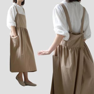 Puede incluir: Un vestido delantal color beige con falda fruncida y grandes bolsillos delanteros. El vestido tiene tirantes anchos y se lleva sobre una blusa blanca con detalles de volantes. La modelo lleva bailarinas beige.