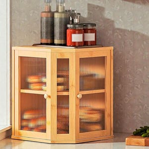 May include: A light-coloured wooden cabinet with glass doors and shelves. The cabinet has a rectangular shape with a slightly angled top. Jars and bottles are on top of the cabinet. The cabinet is likely used for kitchen storage.
