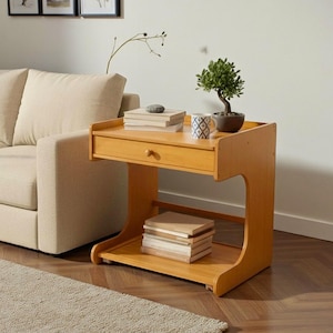 May include: A light brown wooden side table with a drawer and a lower shelf. The table has a curved design on the sides. A stack of books, a small bonsai tree, and a decorative mug are on top. The table is next to a beige sofa.