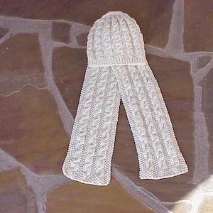 May include: A white knitted hat and scarf set with a cable knit pattern. The hat and scarf are laid out on a brown stone surface.