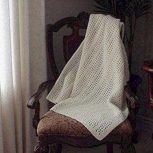 Può includere: Una coperta bianca all'uncinetto drappeggiata su una sedia di legno con un sedile in tessuto a fantasia. La coperta ha un delicato disegno traforato.