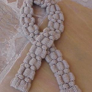 May include: A hand-knitted scarf in a neutral beige color. The scarf features a unique textured design with raised, rounded sections, creating a three-dimensional effect. The scarf is made of a soft, possibly wool or acrylic yarn, and is displayed on a light brown surface.