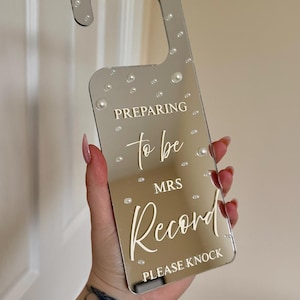 Può includere: Un appendiabiti a specchio con la scritta "Preparing to be Mrs. Record, Please Knock." L'appendiabiti argentato è decorato con piccoli abbellimenti a forma di perla. Un oggetto decorativo per un matrimonio.