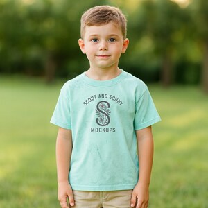 Puede incluir: Un niño lleva una camiseta de color verde azulado claro con el texto "SCOUT AND SONNY MOCKUPS" en un diseño circular. El diseño incluye una "S" floral en el centro. El niño está de pie frente a un fondo verde borroso.