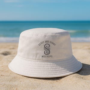May include: A white bucket hat with the text "SCOUT AND SONNY MOCKUPS" in gray. The hat is sitting on a sandy beach with the ocean in the background. The hat has a wide brim and a round crown.