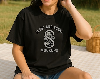 May include: Black t-shirt with the text "SCOUT AND SONNY MOCKUPS" in a silver color. The design features a stylized "S" with floral accents. The model is wearing denim shorts and is sitting on a picnic blanket.