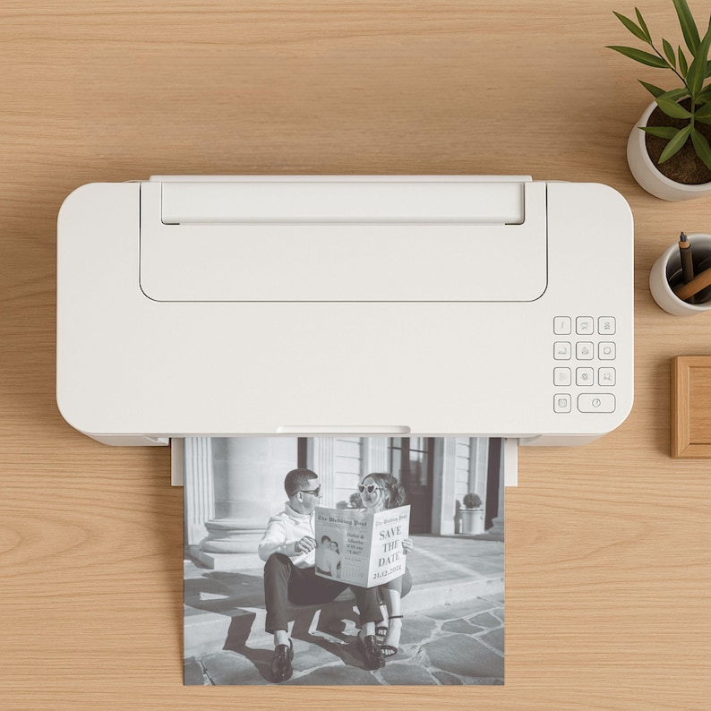 May include: A white printer is printing a black and white photo of a couple holding a newspaper with the text "SAVE THE DATE 20.03.2019". The printer is on a light brown wooden surface, with a small plant and pencils in the background.