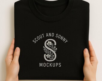 May include: A black sweatshirt with a crew neck, held by two hands. The sweatshirt features a white floral design with the text "SCOUT AND SONNY MOCKUPS" in a circular arrangement above and below the floral design.