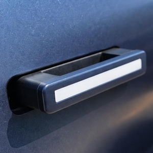 May include: Close-up of a dark blue car door with a black door handle. The handle has a rectangular shape with a white accent stripe. The car door has a smooth, metallic finish.