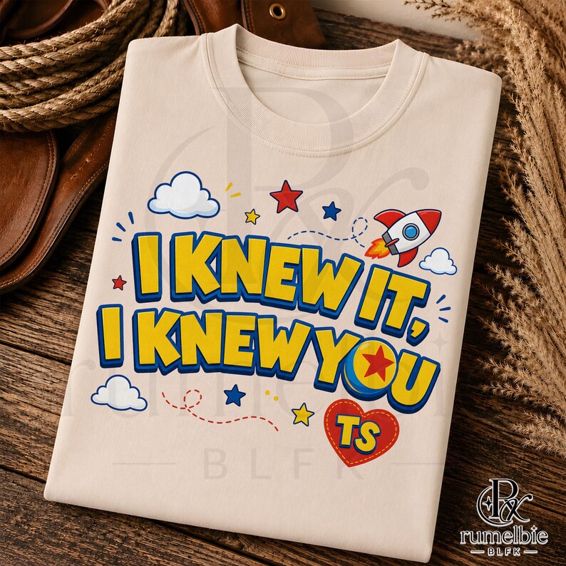 May include: A beige t-shirt featuring the text I KNEW IT, I KNEW YOU in bold yellow letters with blue outlines. The design includes a red and white rocket, white clouds, colorful stars, and a red heart containing the yellow letters TS.