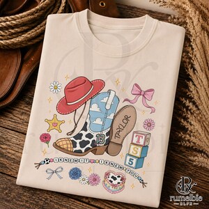 May include: A cream-colored t-shirt featuring a graphic print of a red cowboy hat, a cow-print boot with a blue sky and cloud pattern, the number 13, the name Taylor, a pink bow, star, flowers, blocks labeled T, S, 5, and a beaded string with text.