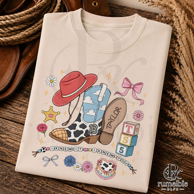 May include: A cream-colored t-shirt featuring a graphic print of a red cowboy hat, a cow-print boot with a blue sky and cloud pattern, the number 13, the name Taylor, a pink bow, star, flowers, blocks labeled T, S, 5, and a beaded string with text.