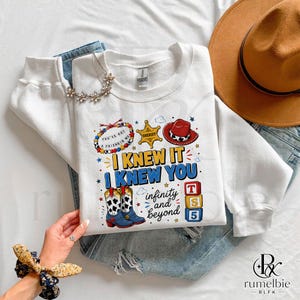May include: A white long-sleeve sweatshirt featuring a graphic print with a sheriff badge, a red cowboy hat, cow-print boots, and colorful blocks. The text reads I KNEW IT I KNEW YOU infinity and beyond, with a beaded bracelet graphic saying YOU'VE GOT A FRIEND IN ME.