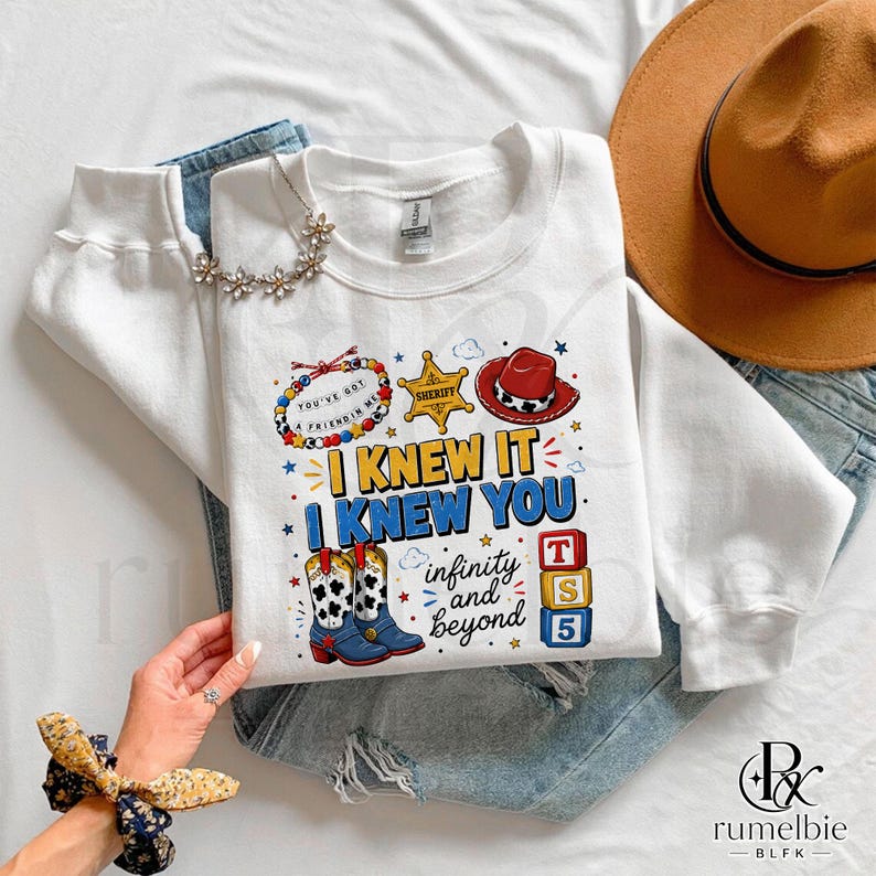 May include: A white long-sleeve sweatshirt featuring a graphic print with a sheriff badge, a red cowboy hat, cow-print boots, and colorful blocks. The text reads I KNEW IT I KNEW YOU infinity and beyond, with a beaded bracelet graphic saying YOU'VE GOT A FRIEND IN ME.
