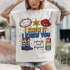 May include: A cream-colored short-sleeve t-shirt featuring a graphic print with a beaded bracelet, a yellow sheriff badge, a red cowboy hat, and blue cowboy boots. The text reads I KNEW IT I KNEW YOU, infinity and beyond, and T S 5 on blocks.