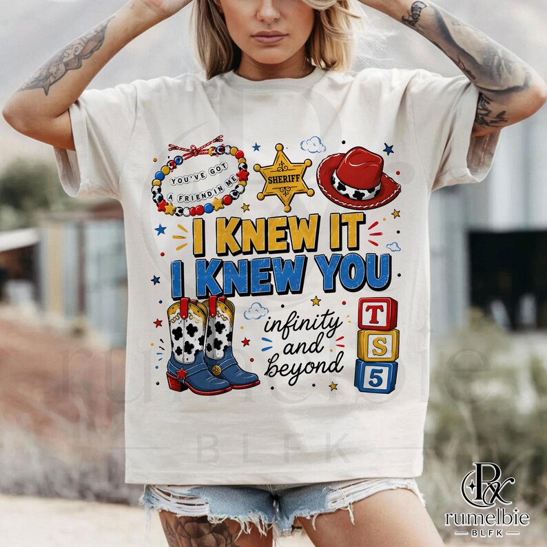 May include: A cream-colored short-sleeve t-shirt featuring a graphic print with a beaded bracelet, a yellow sheriff badge, a red cowboy hat, and blue cowboy boots. The text reads I KNEW IT I KNEW YOU, infinity and beyond, and T S 5 on blocks.