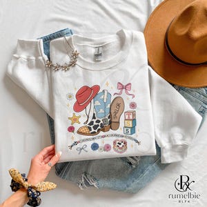 May include: A white crewneck sweatshirt featuring a graphic print of a cowboy boot with a cow print pattern, a red cowboy hat, a pink bow, building blocks labeled T, S, 5, and the text "I KNEW IT I KNEW YOU" with the name "TAYLOR" on the boot sole.