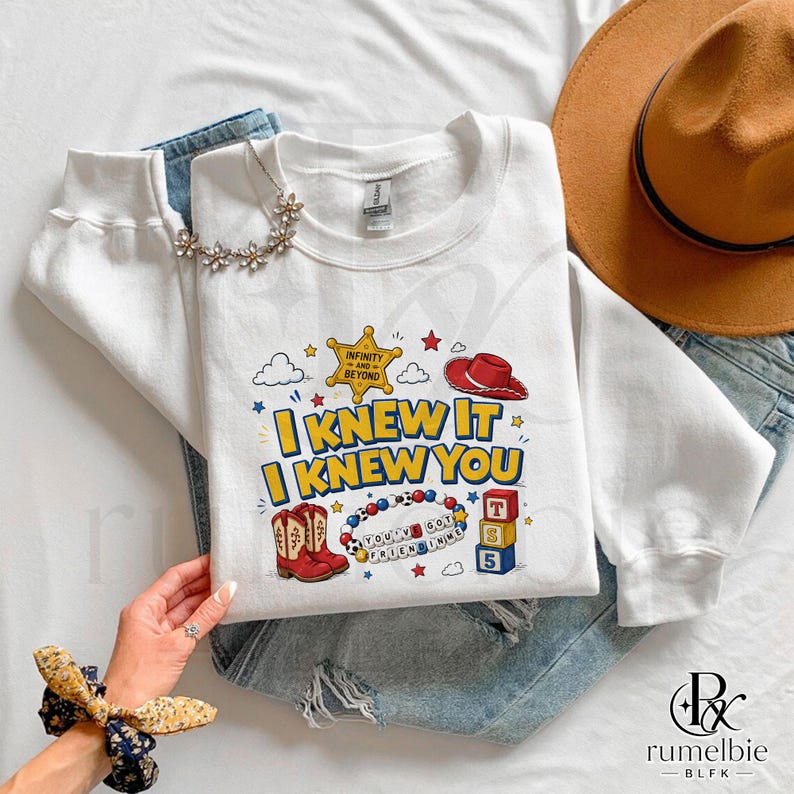 May include: A white long-sleeve sweatshirt featuring a graphic print with the text I KNEW IT I KNEW YOU, a yellow sheriff badge with the text INFINITY AND BEYOND, a red cowboy hat, red cowboy boots, a beaded bracelet, and three stacked alphabet blocks.