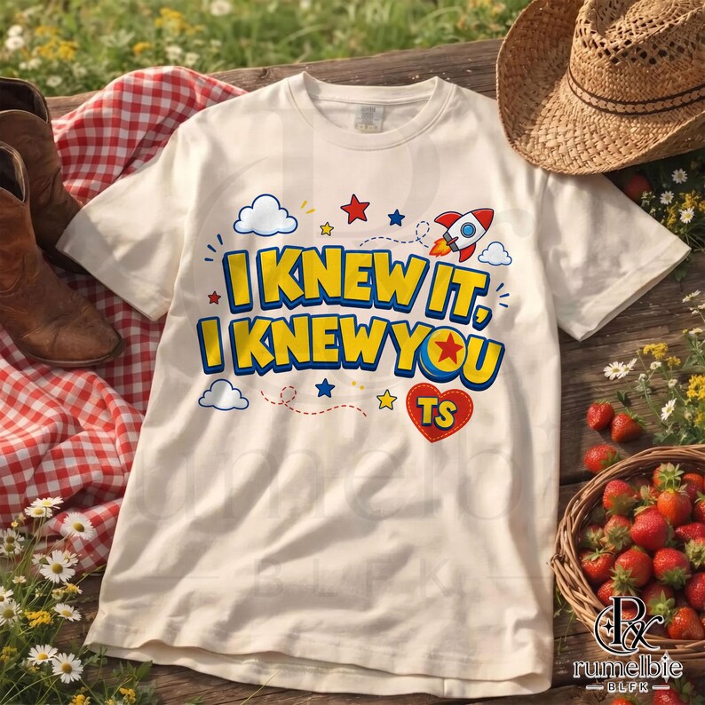 May include: A cream-colored t-shirt featuring the text I KNEW IT, I KNEW YOU in bold blue and yellow letters. The design includes a small rocket ship, clouds, stars, and a red heart with the letters TS inside, all printed on the front of the shirt.