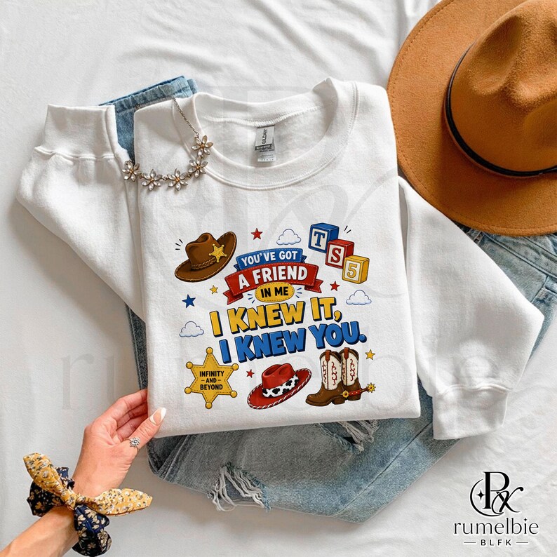 Puede incluir: Una sudadera blanca de manga larga con un estampado gr&aacute;fico que muestra un sombrero de vaquero marr&oacute;n, una estrella de sheriff amarilla, un sombrero de vaquero rojo y bloques. El texto dice: You've got a friend in me, I knew it, I knew you.