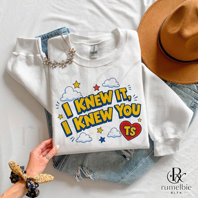 May include: A white long-sleeve sweatshirt featuring the text I KNEW IT, I KNEW YOU in yellow with blue outlines. The design includes blue and white clouds, small red, yellow, and blue stars, and a red heart with the letters TS inside.