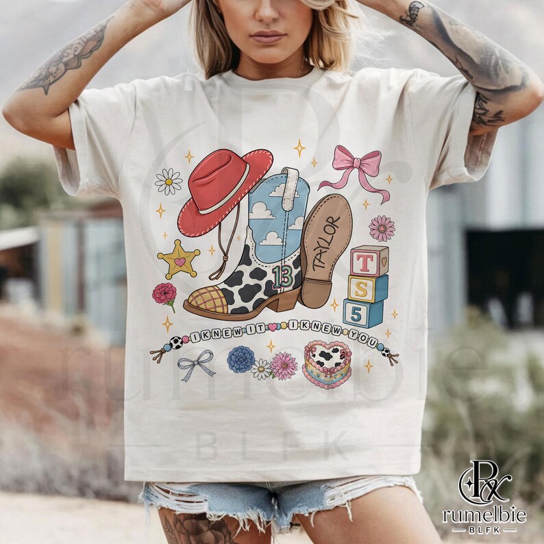 May include: A cream-colored t-shirt featuring a graphic print of a red cowboy hat, a cow-print boot with the number 13, a pink bow, alphabet blocks spelling T S 5, a heart-shaped cake, and the text I KNEW IT I KNEW YOU with the name TAYLOR.