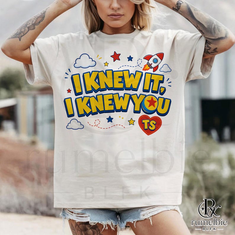 May include: A cream-colored short-sleeve t-shirt featuring the text I KNEW IT, I KNEW YOU in bold yellow letters with blue outlines. The design includes a red rocket, blue and red stars, white clouds, and a red heart with the letters TS inside.