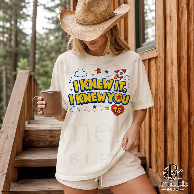 May include: A cream-colored short-sleeve t-shirt featuring the text I KNEW IT, I KNEW YOU in blue and yellow letters. The design includes a red heart with TS, a small rocket ship, blue clouds, and red and yellow stars on the front of the shirt.