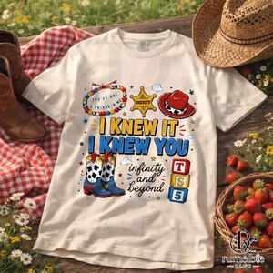 May include: A cream-colored t-shirt featuring a graphic print with the text I KNEW IT I KNEW YOU and infinity and beyond. The design includes a beaded bracelet, a gold sheriff badge, a red cowboy hat, two cow-print boots, and three toy blocks.
