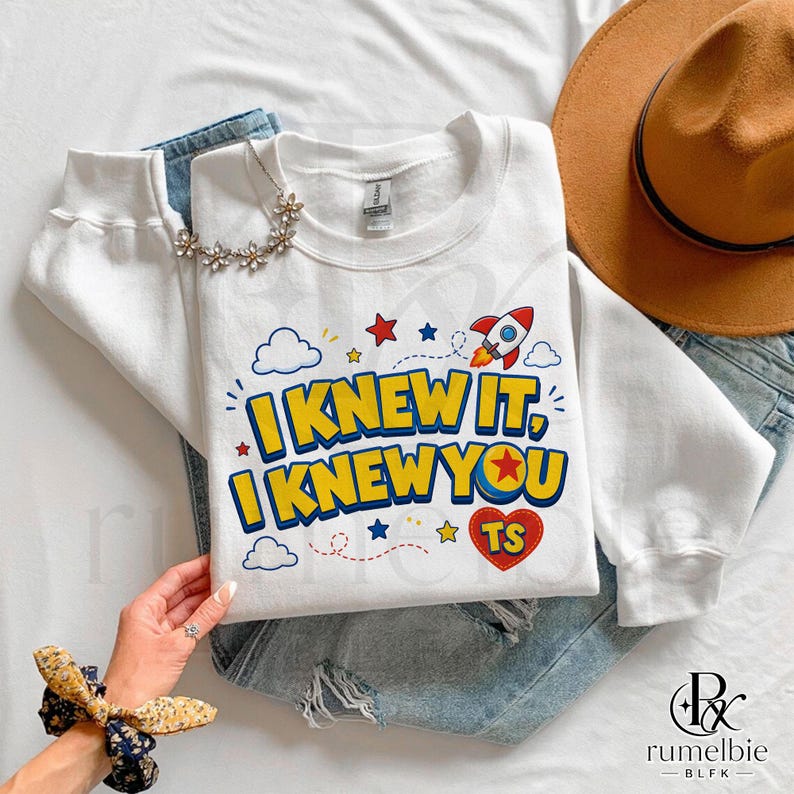 May include: A white long-sleeve sweatshirt featuring a graphic print with the text I KNEW IT, I KNEW YOU and TS inside a red heart. The design includes a rocket ship, blue clouds, and yellow and red stars. The sweatshirt is layered over blue denim jeans.