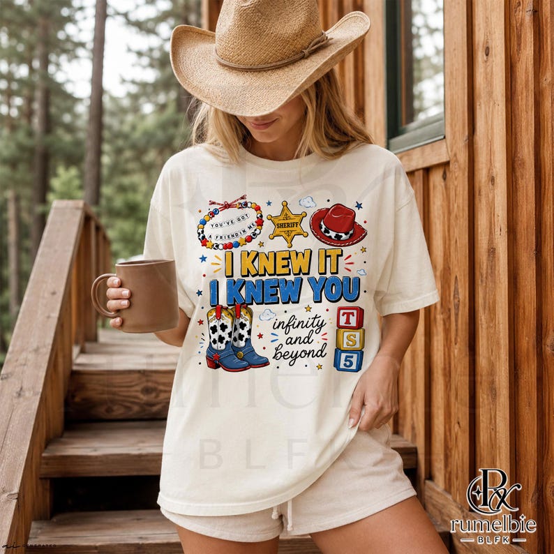 May include: A cream-colored short-sleeve t-shirt featuring a graphic print with the text I KNEW IT I KNEW YOU infinity and beyond. The design includes a beaded bracelet, a sheriff star, a red cowboy hat, cow-print boots, and three alphabet blocks.